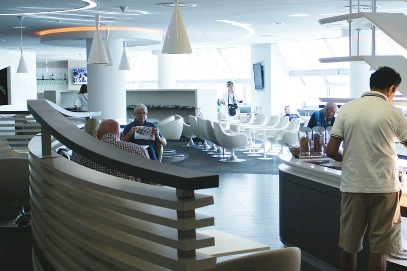 Helle Flughafenlounge mit modernen Sitzgruppen, Buffetinsel rechts und Gästen beim Lesen und Essen
