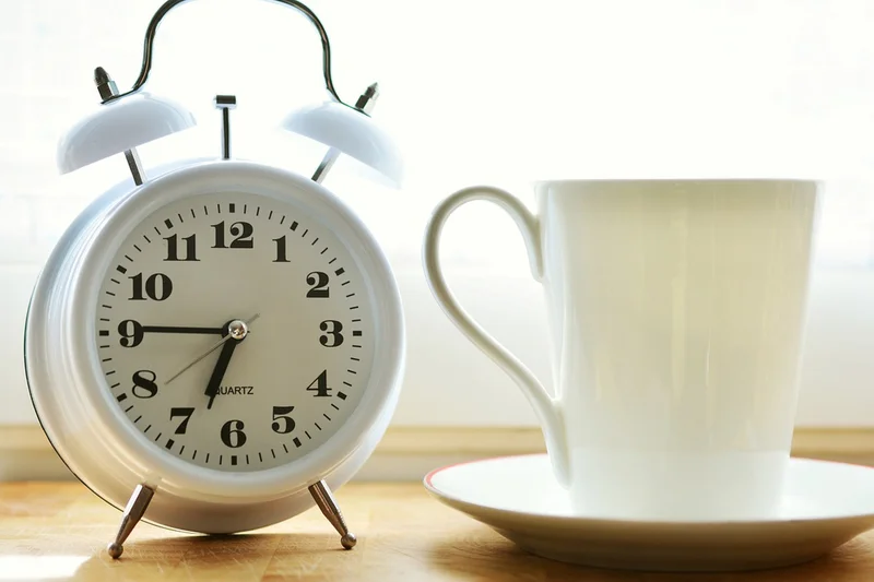 Weißer Doppelglockenwecker auf Holzfläche zeigt Zeit neben großer weißer Tasse auf Untertasse am Fenster