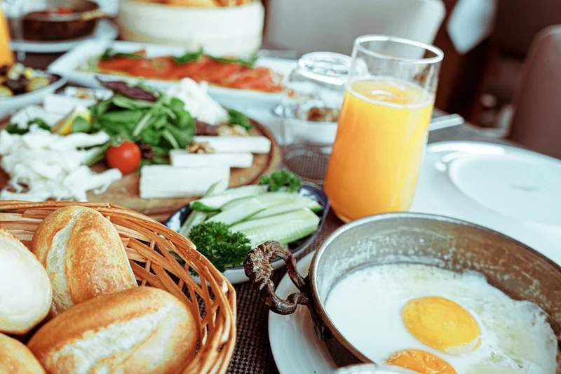 Brotkorb mit Brötchen, Pfanne mit Spiegeleiern, Glas Orangensaft und Schalen mit Gurken, Käse und Salat