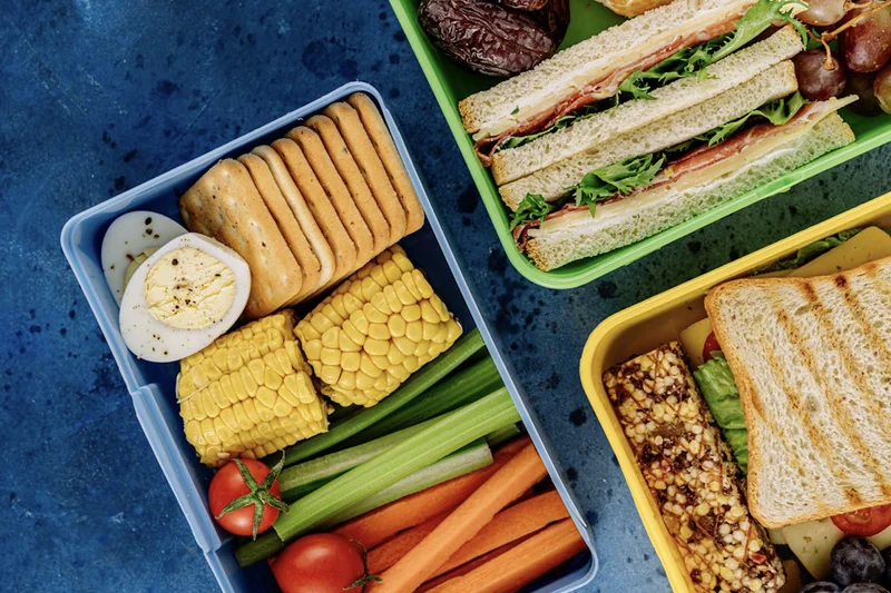 Drei gefüllte Brotdosen mit Sandwiches, hartgekochtem Ei, Crackern, Mais, Rohkost und Müsliriegeln